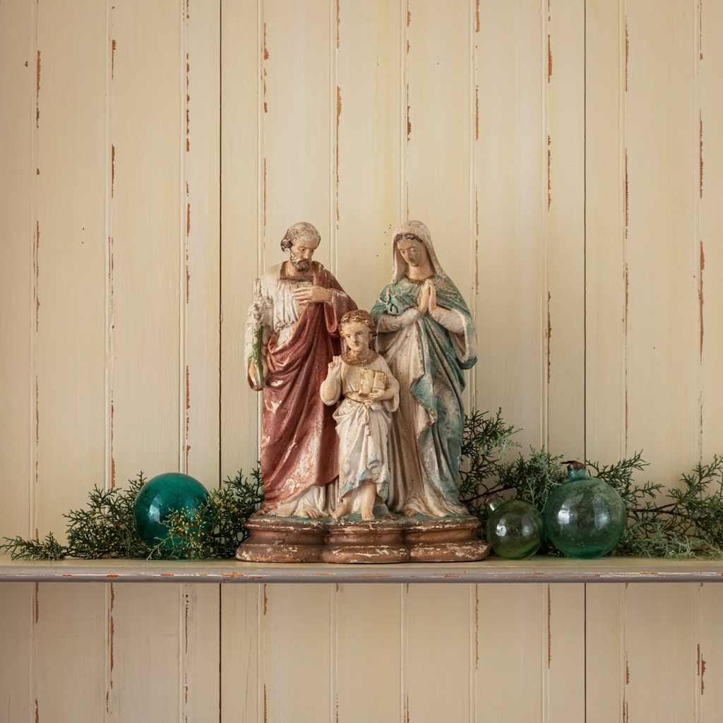 Statue of the Holy Family- Joseph, Mary, and Jesus- on a shelf with seasonal decorative elements against a wooden paneled wall.