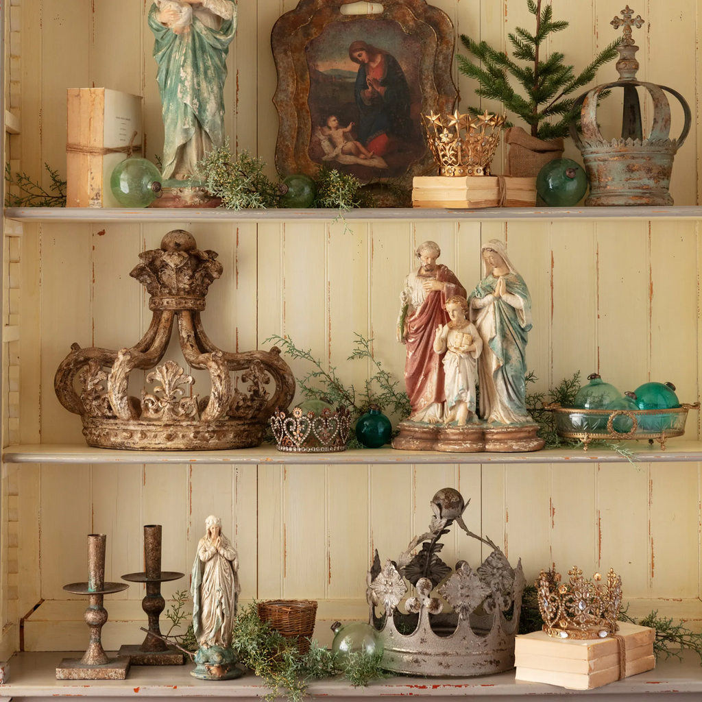 Decorative shelves with vintage figurines such as the Holy Family, crowns, and plants against a wooden wall.