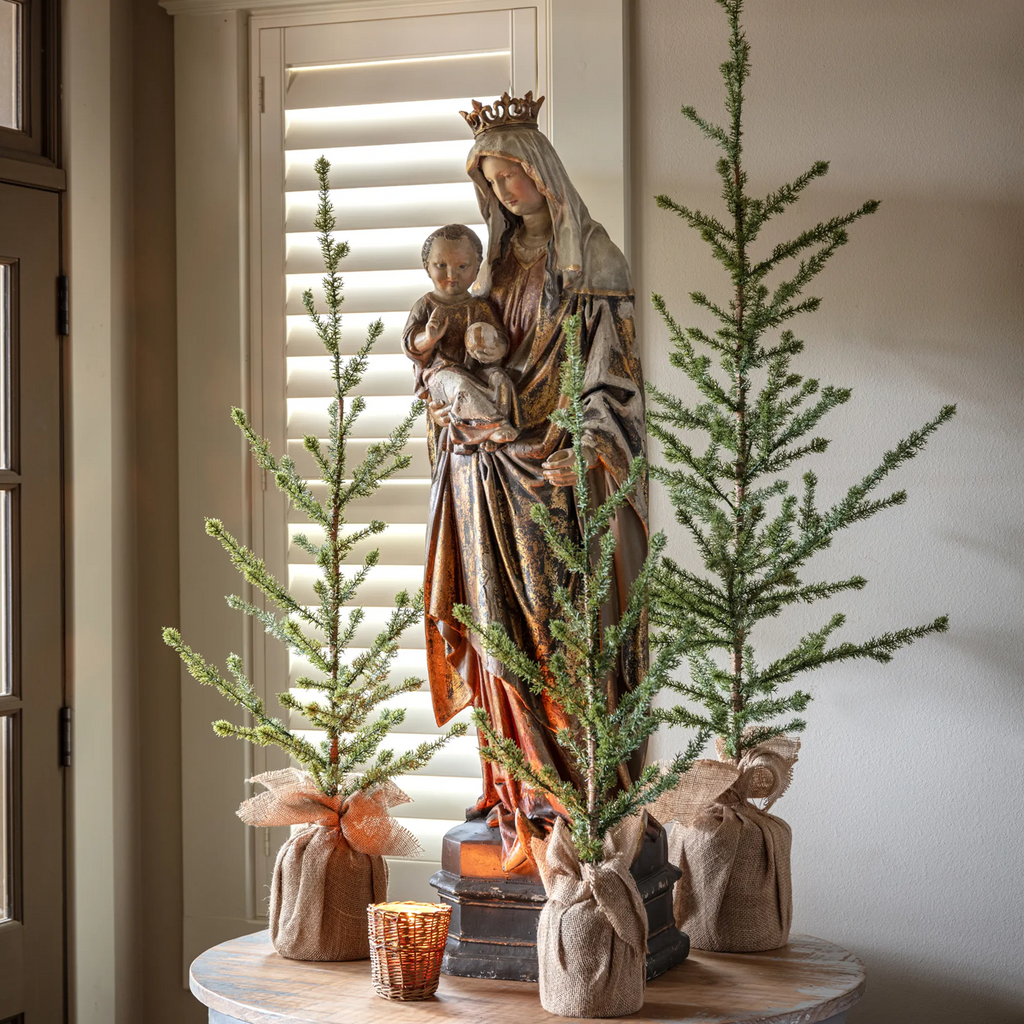 Statue of Mother Mary holding Baby Jesus on a table surrounded by 3 Christmas trees in burlap bags and a candle.