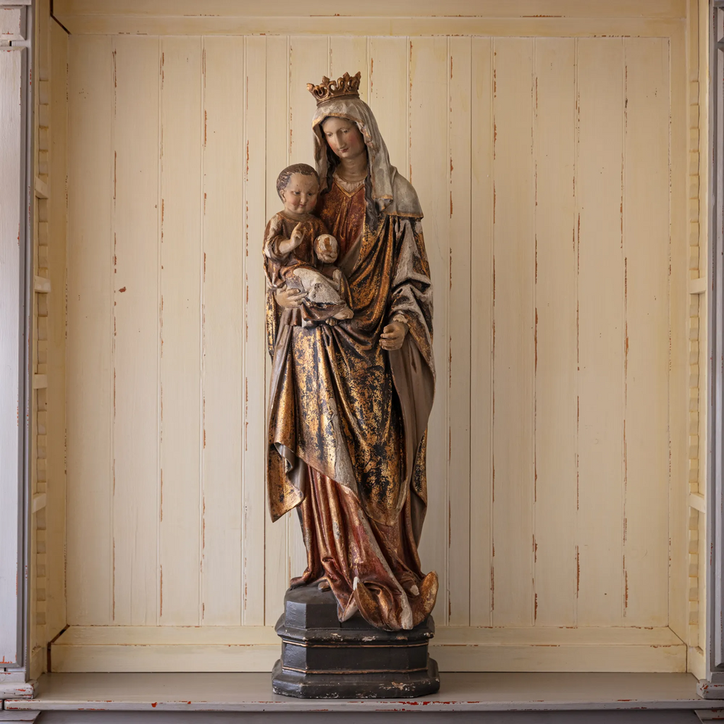 Statue of Mother Mary holding Baby Jesus with a wooden paneled wall in the background.