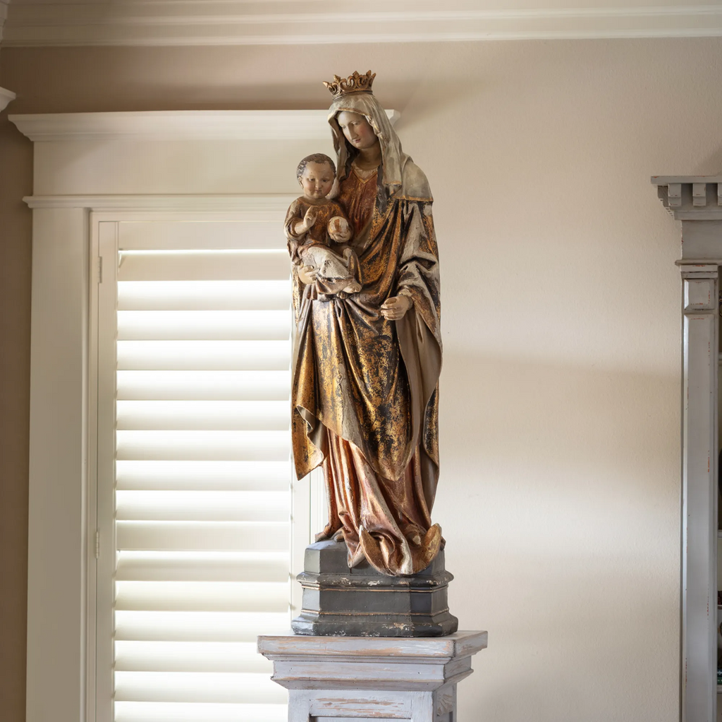 Statue of Mother Mary holding Baby Jesus on a pedestal against a neutral wall.
